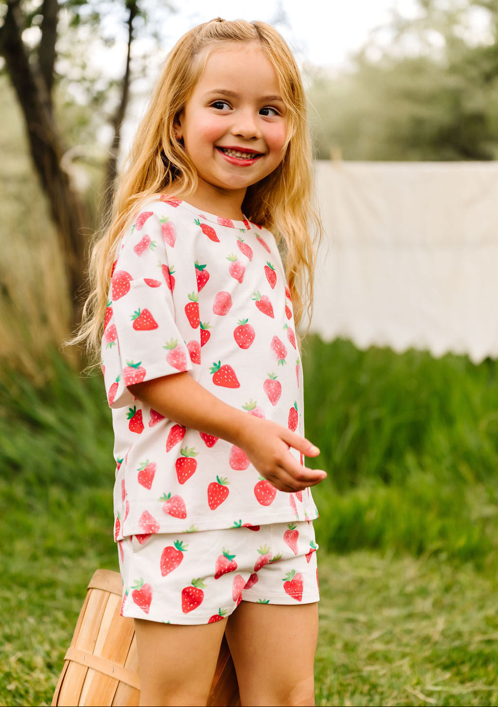 Young girl wearing a strawberry-patterned outfit standing outdoors.